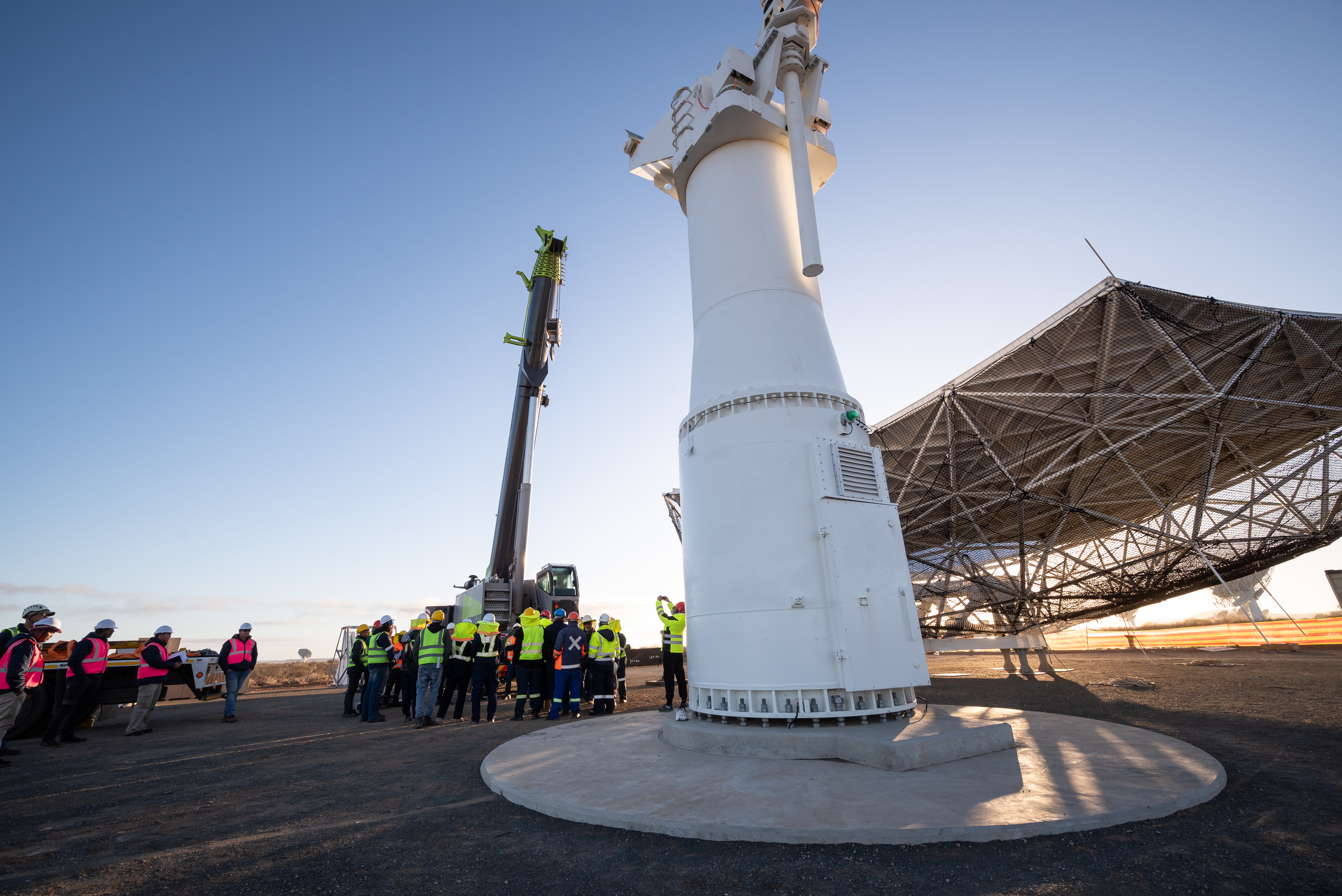 The team assembled under the pedestal and a crane as dawn breaks on site. To their right, the huge main reflected sits on the ground.