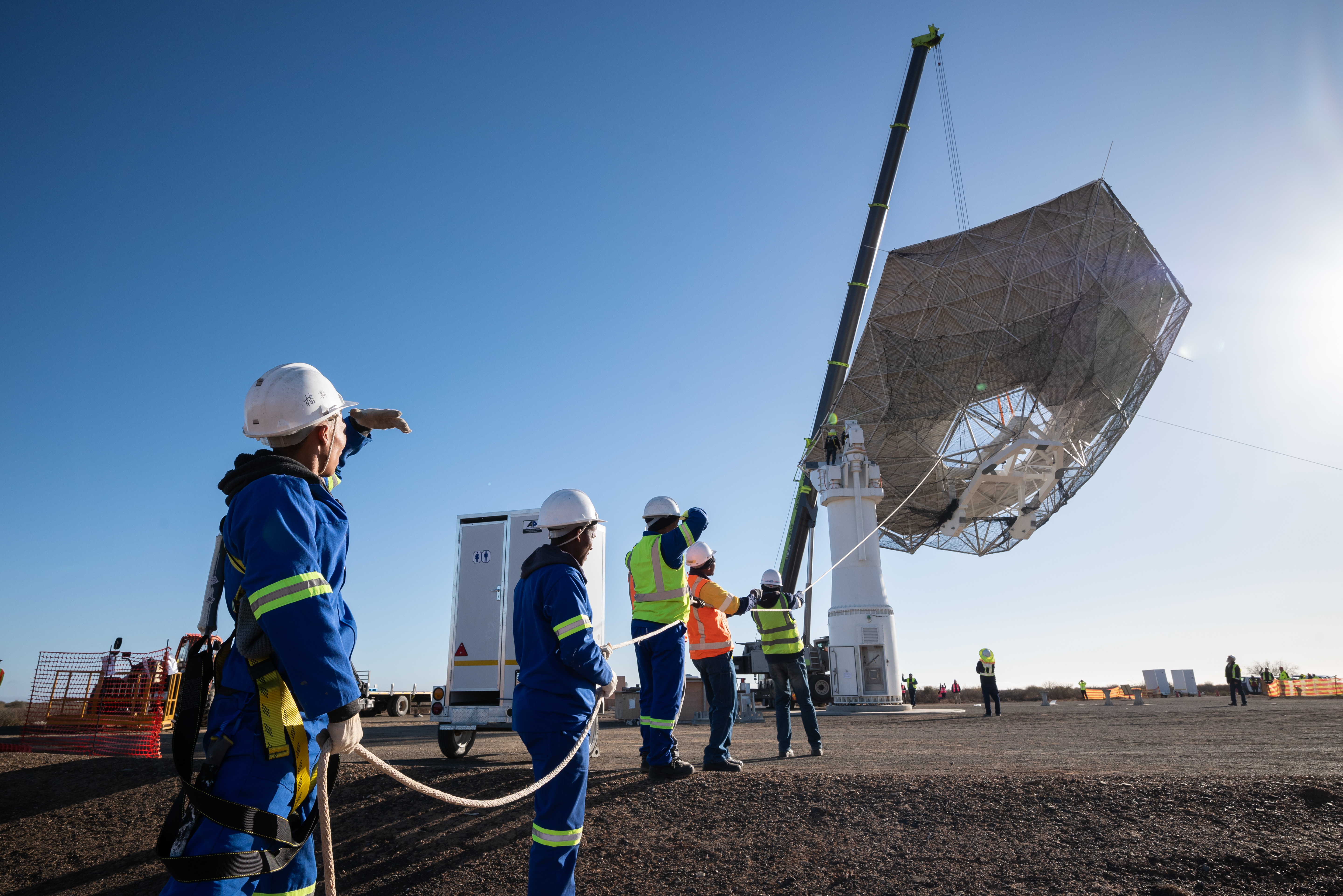A team holding a rope as the huge main reflector is raised into the air by a crane.