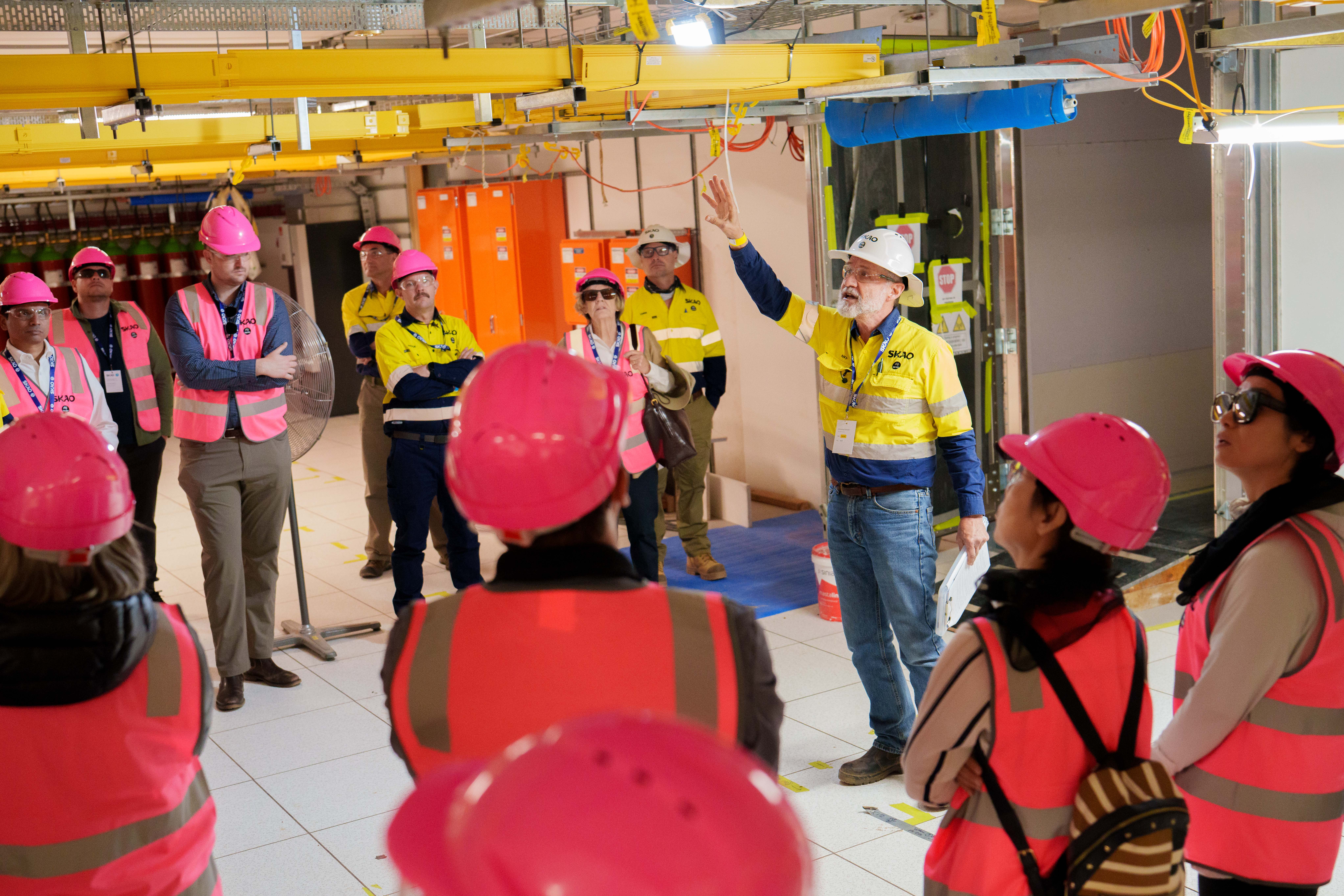 A man in a yellow hi-vis shirt and hard hat surrounded by people in pink hi-vis vests and hard hats. He is explaining something and pointing to the ceiling of the room they are in.