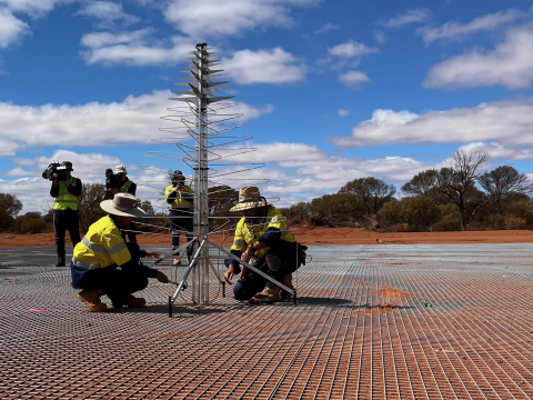 The first SKA-Low antenna being installed on its ground mesh by two technicians. In the background, a camera crew films the moment.