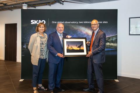 Photo of three people, from left: STFC Executive Chair Michele Dougherty, SKAO Director-General Prof. Philip Diamond, and UK Minister of State for Science, Research and Innovation Lord Patrick Vallance. Prof. Diamond is handing Lord Vallance a frame photograph of the SKA telescopes. In the background is an SKAO banner.