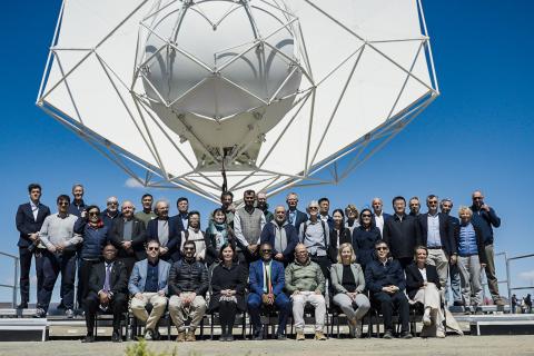 Representatives of 26 countries stand underneath an SKA dish facing the camera under a blue sky.