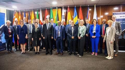 Representatives from the SKA Observatory member states and observers stand in a room smiling in front of flags representing their countries. 