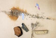 Composition of different elements: A line of sand from left to right with two people next to it. To the bottom are three differently coloured rocks and a tree represented by a twig. 