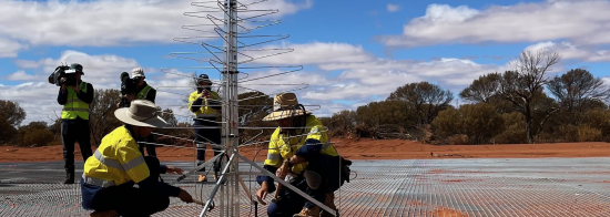 The first SKA-Low antenna being installed on its ground mesh by two technicians. In the background, a camera crew films the moment.