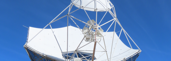 Picture of the first SKA-Mid dish against a blue sky. The dish has a hexagonal shape and sits atop a white pedestal. A smaller spherical shaped mirror is supported by small white bars.  A triangular structure, the feed indexer, is also mounted on these bars.