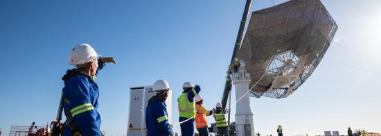 A team holding a rope as the huge main reflector is raised into the air by a crane.