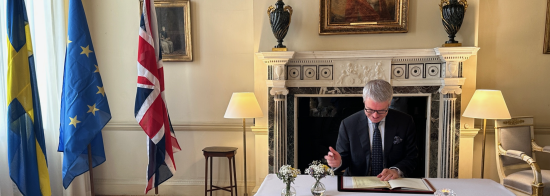 Swedish Ambassador Stefan Gullgren signing the SKAO Convention. He is sitting at a table in front of a fireplace. In the background are the Swedish, EU and UK flags.