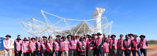 Group picture of group of people in pink high-vis jackets in front of a telescope dish under construction.