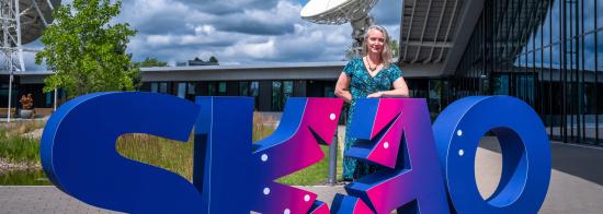 Picture of Prof. Jessica Dempsey standing behind big letters that read "SKAO" in front of the SKAO Global Headquartes.