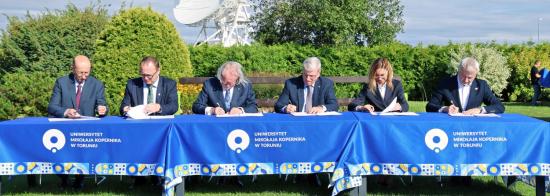 Five men and a woman signing documents on a long table in front of a telescope dish in the background.