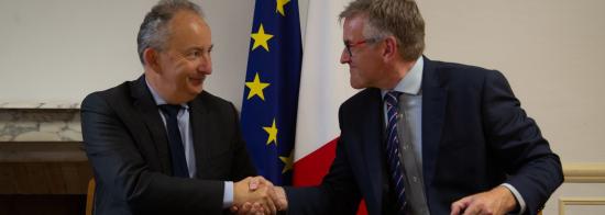 Two men in suits shaking hands, with a document in front of them. In the background are the EU and French flags.