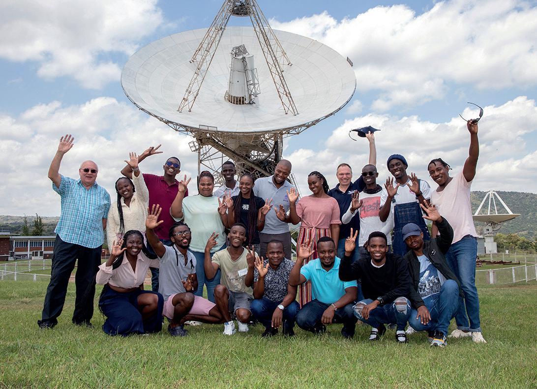 DARA students from Kenya and South Africa wave in front of a radio telescope at SARAO Hartebeesthoek Radio Astronomy Observatory.