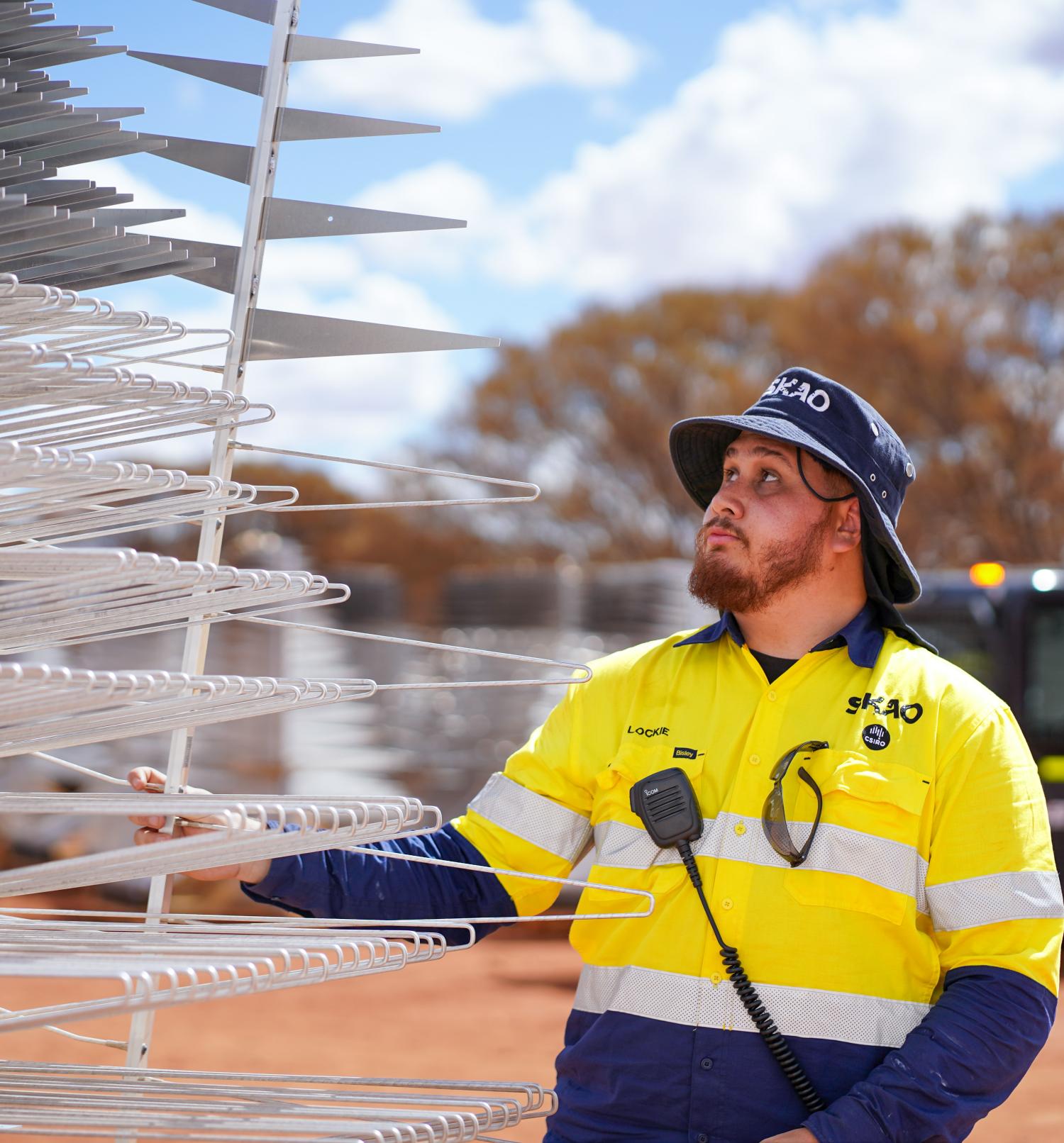A Wajarri SKA-Low field technician wearing a hi-vis shirt and SKAO hat, lifting an SKA-Low antenna.