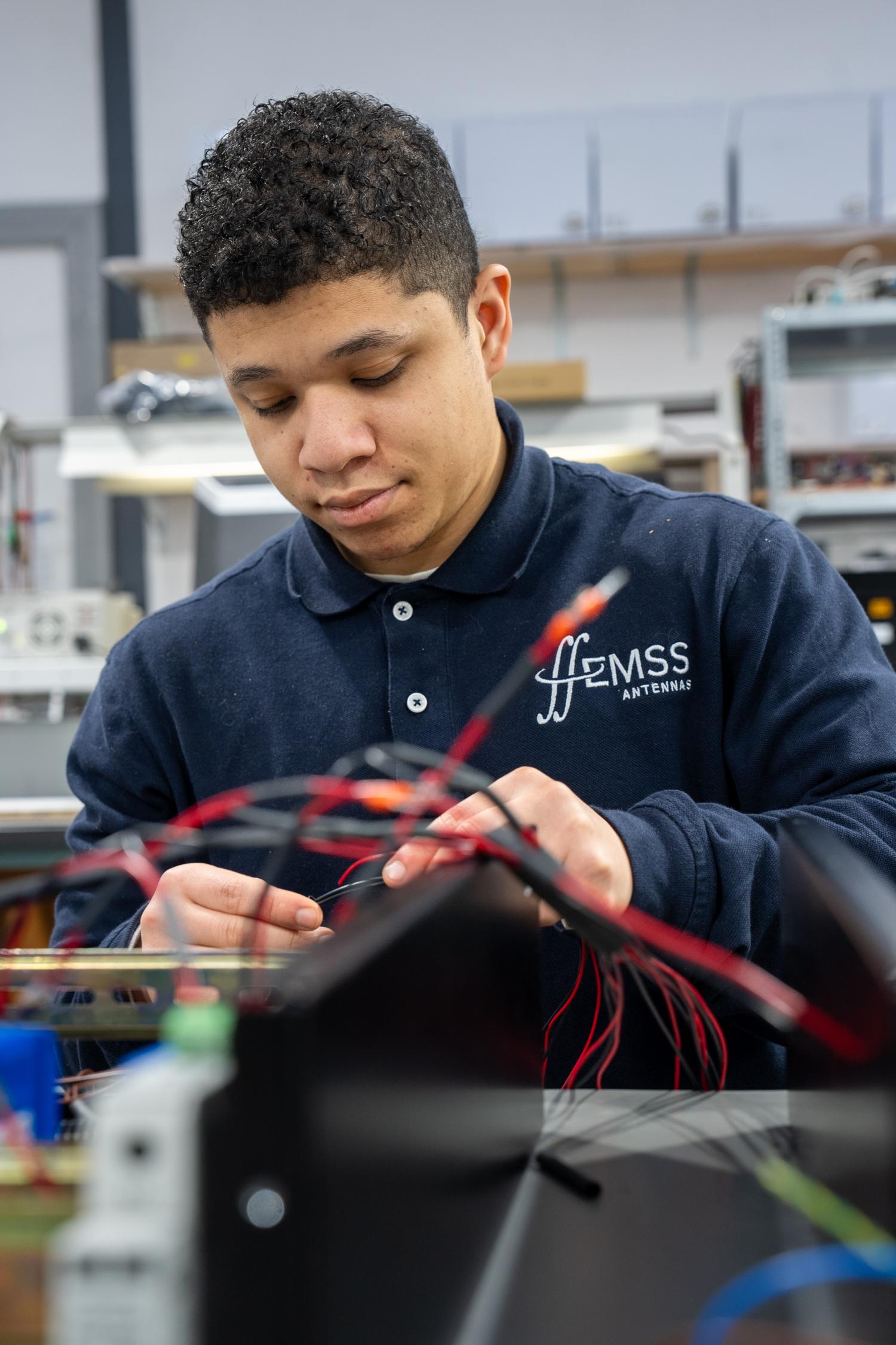 A man working with electrical wires in a facility. The employee is wearing a blue shirt with the EMSS Antennas logo.