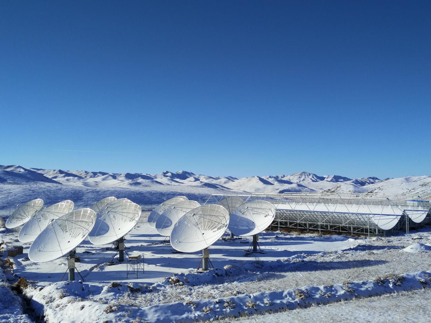 Nine telescope dishes and three open cylindrical telescopes in a snowy mountainous landscape under clear blue skies.
