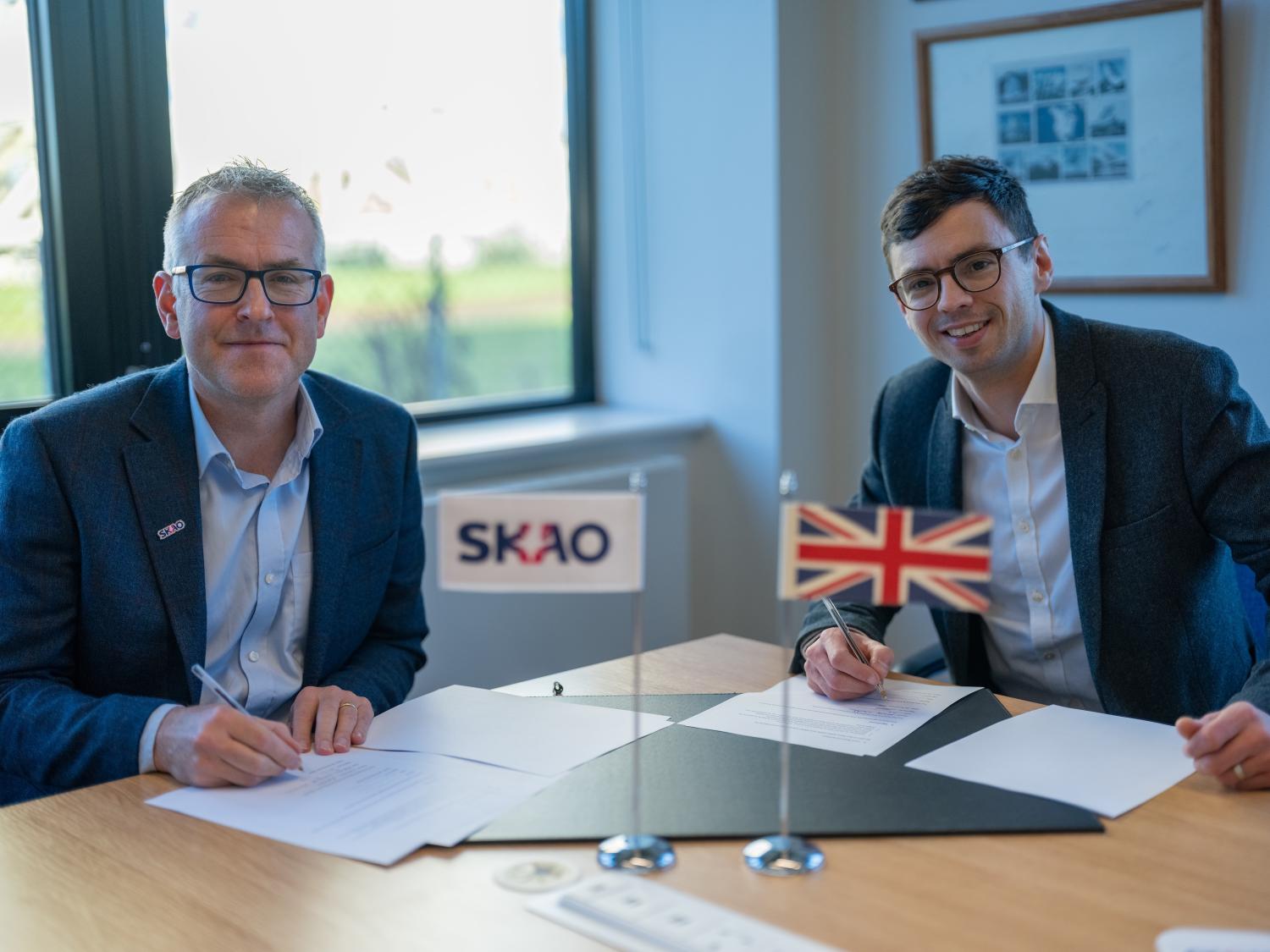 Two men signing documents. In the front are two flags, one shows the SKAO logo and the other is the UK flag.