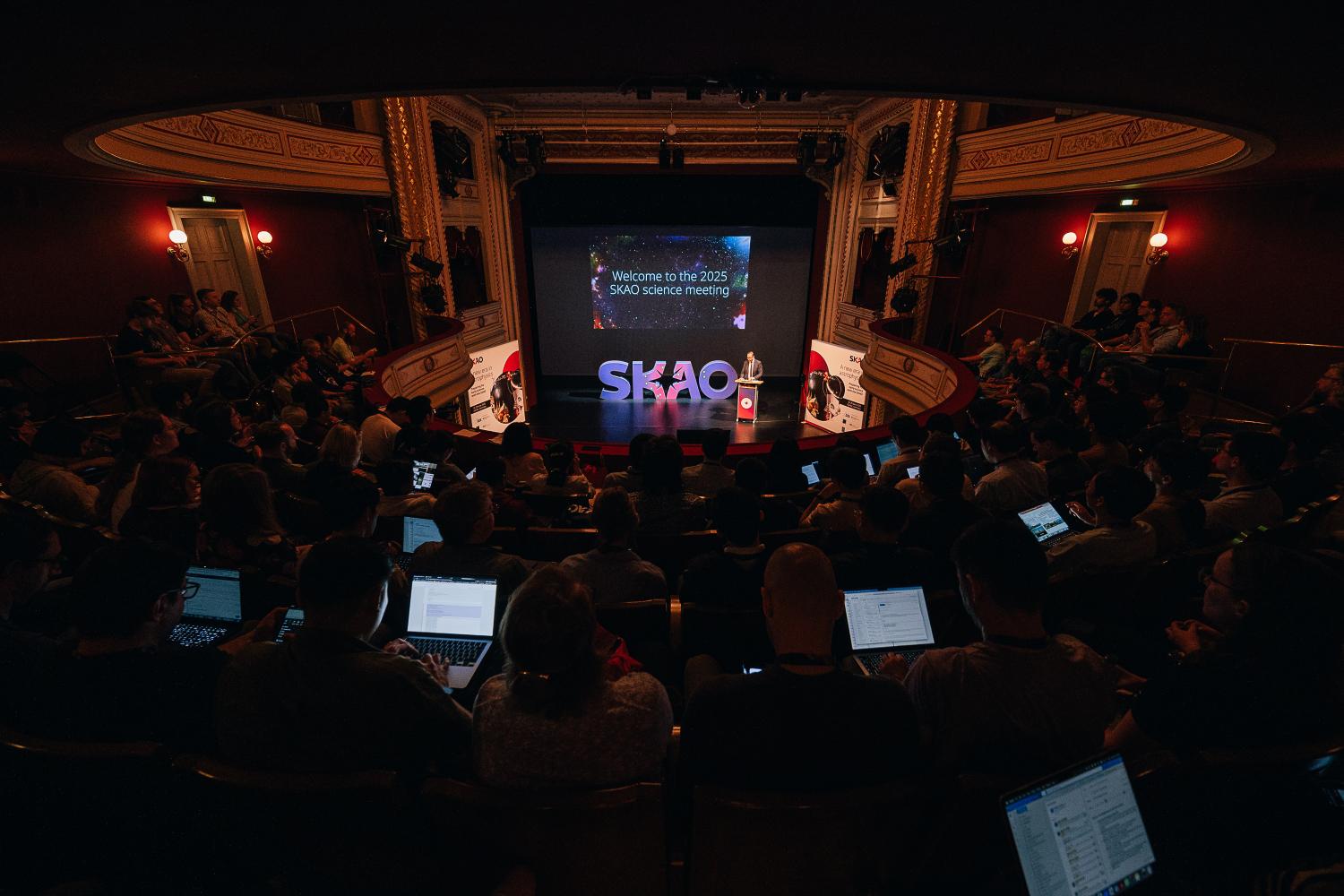 A view from the top of the auditorium during the SKAO science meeting, with people on laptops, and the stage in the background with the SKAO logo