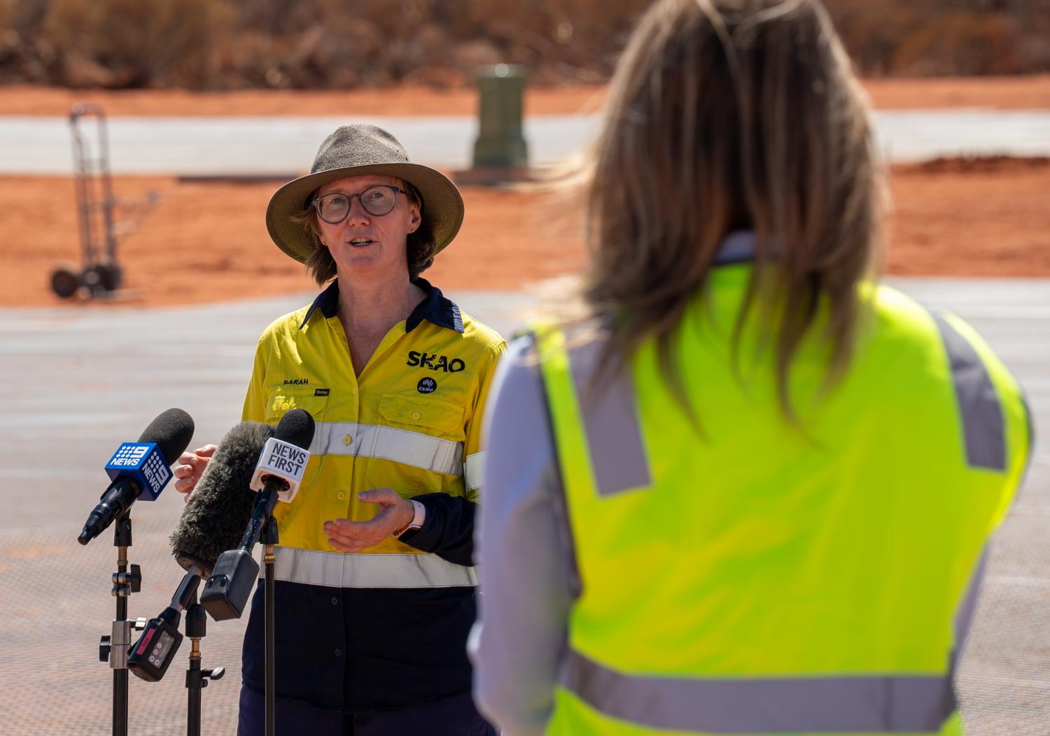 a woman wearing a hivis jacket with the SKAO and CSIRO logos speaks to a group of media microphones on the SKA-Low site in Australia.