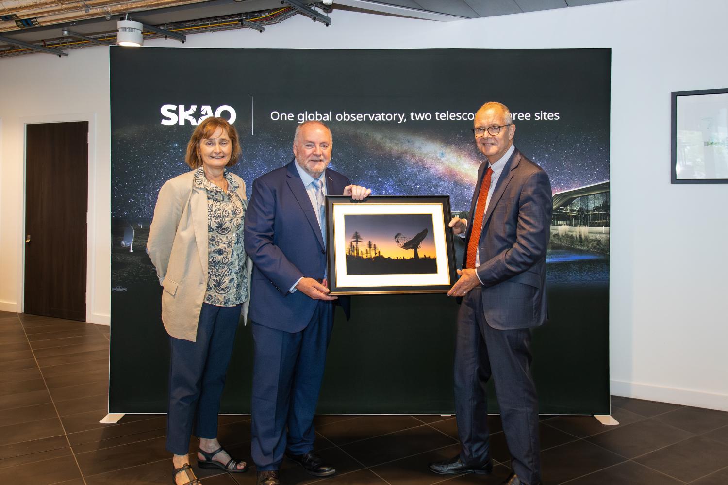 Photo of three people, from left: STFC Executive Chair Michele Dougherty, SKAO Director-General Prof. Philip Diamond, and UK Minister of State for Science, Research and Innovation Lord Patrick Vallance. Prof. Diamond is handing Lord Vallance a frame photograph of the SKA telescopes. In the background is an SKAO banner.