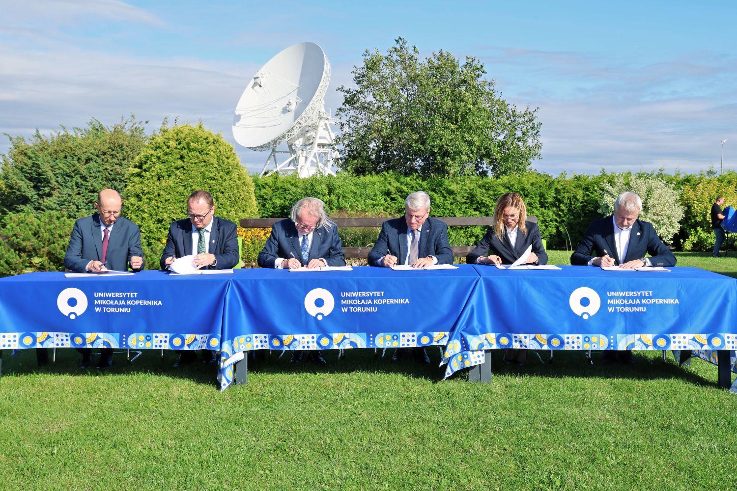 Five men and a woman signing documents on a long table in front of a telescope dish in the background.