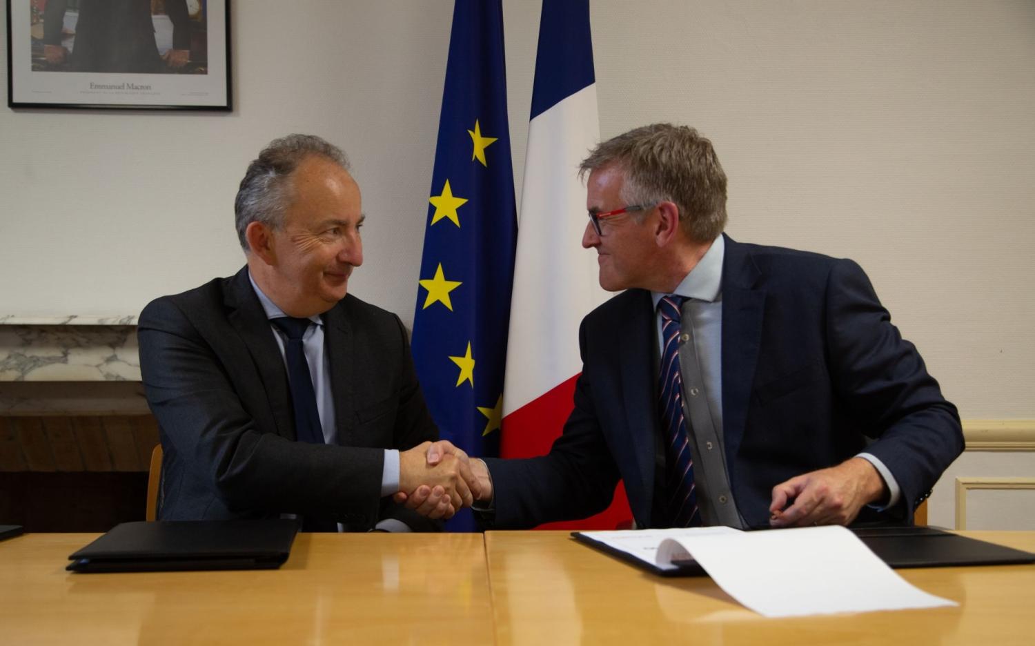 Two men in suits shaking hands, with a document in front of them. In the background are the EU and French flags.