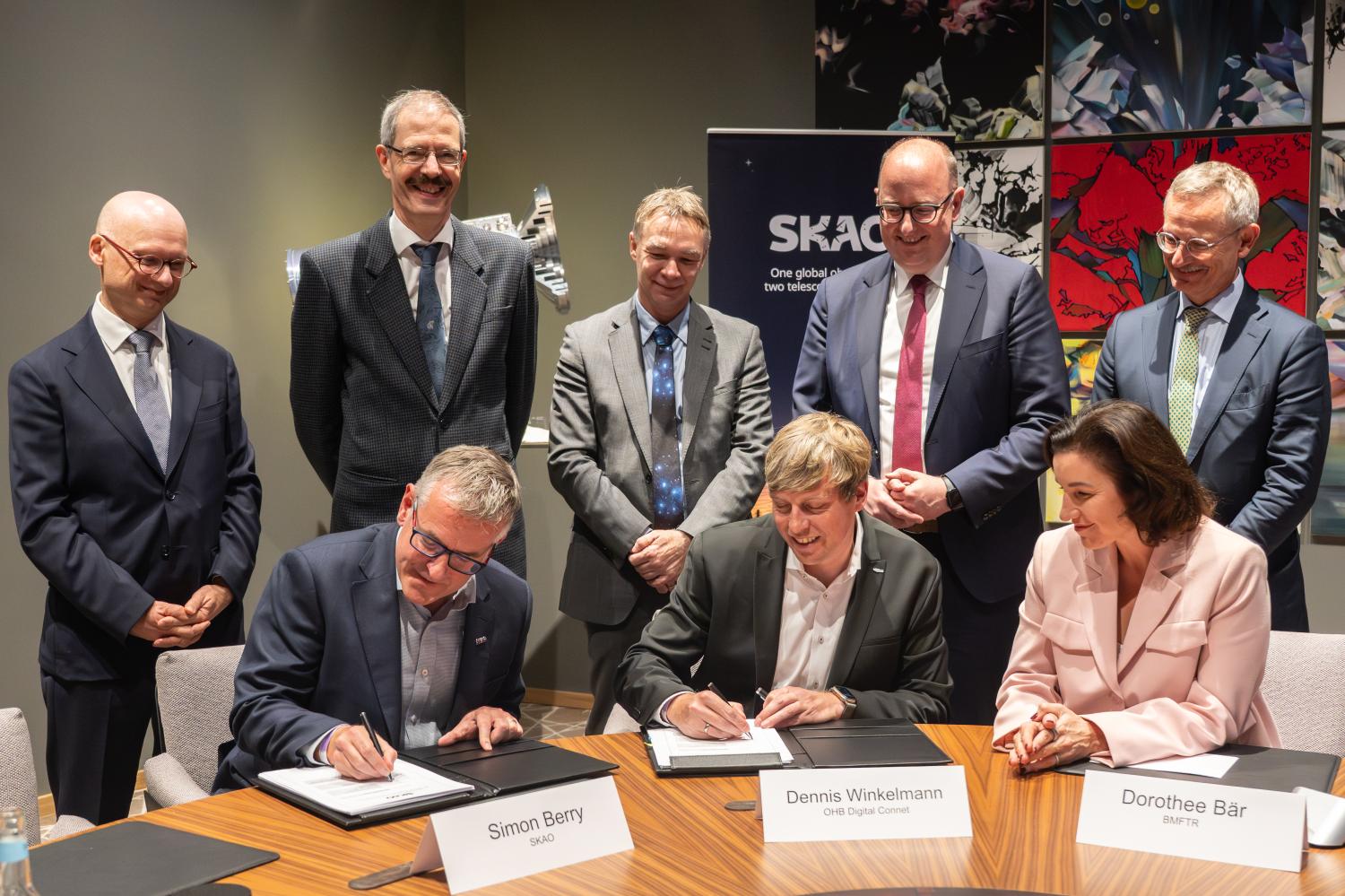 SKAO Deputy DG Simon Berry and OHB Managing Director Dennis Winckelmann sign a contract as six others look on with encouraging smiles.