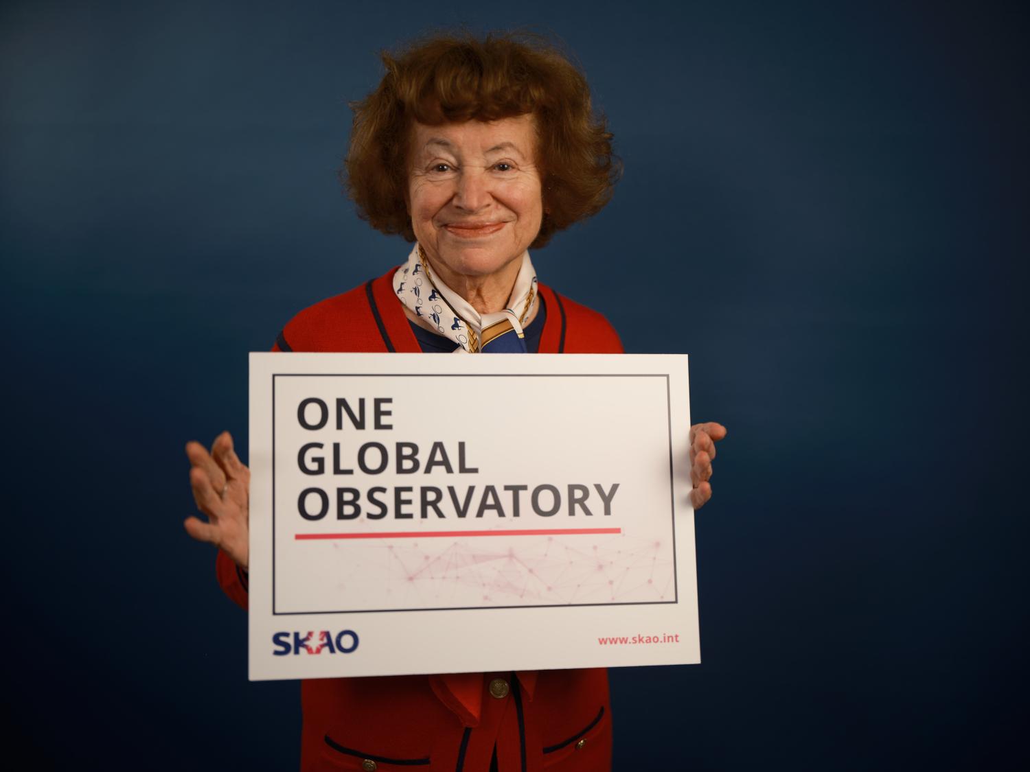 Dr Catherine Cesarsky holding a sign that reads "One global observatory - SKAO"